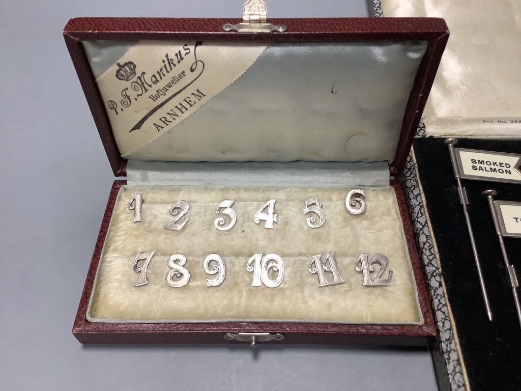 A cased set of twelve Dutch white metal number place settings, 12mm and a cased set of six silver sandwich flags sticks, height 8cm.